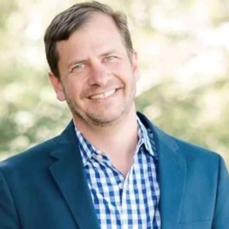 The image shows a man with short brown hair and a friendly smile. He is wearing a blue blazer over a blue and white checkered shirt. The background is blurred, suggesting an outdoor setting. The man appears to be in his late 40s or early 50s.